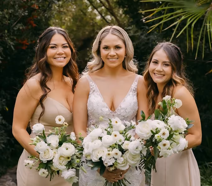 A bride and two bridesmaids holding crisp white flowers enveloped by green foliage