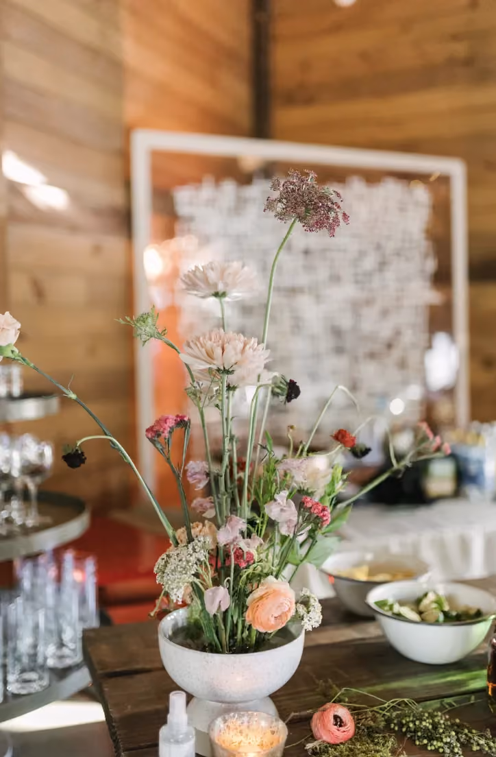 Ikebana-inspired floral arrangement with delicate wildflowers in a white bowl on a rustic wooden table at a wedding.