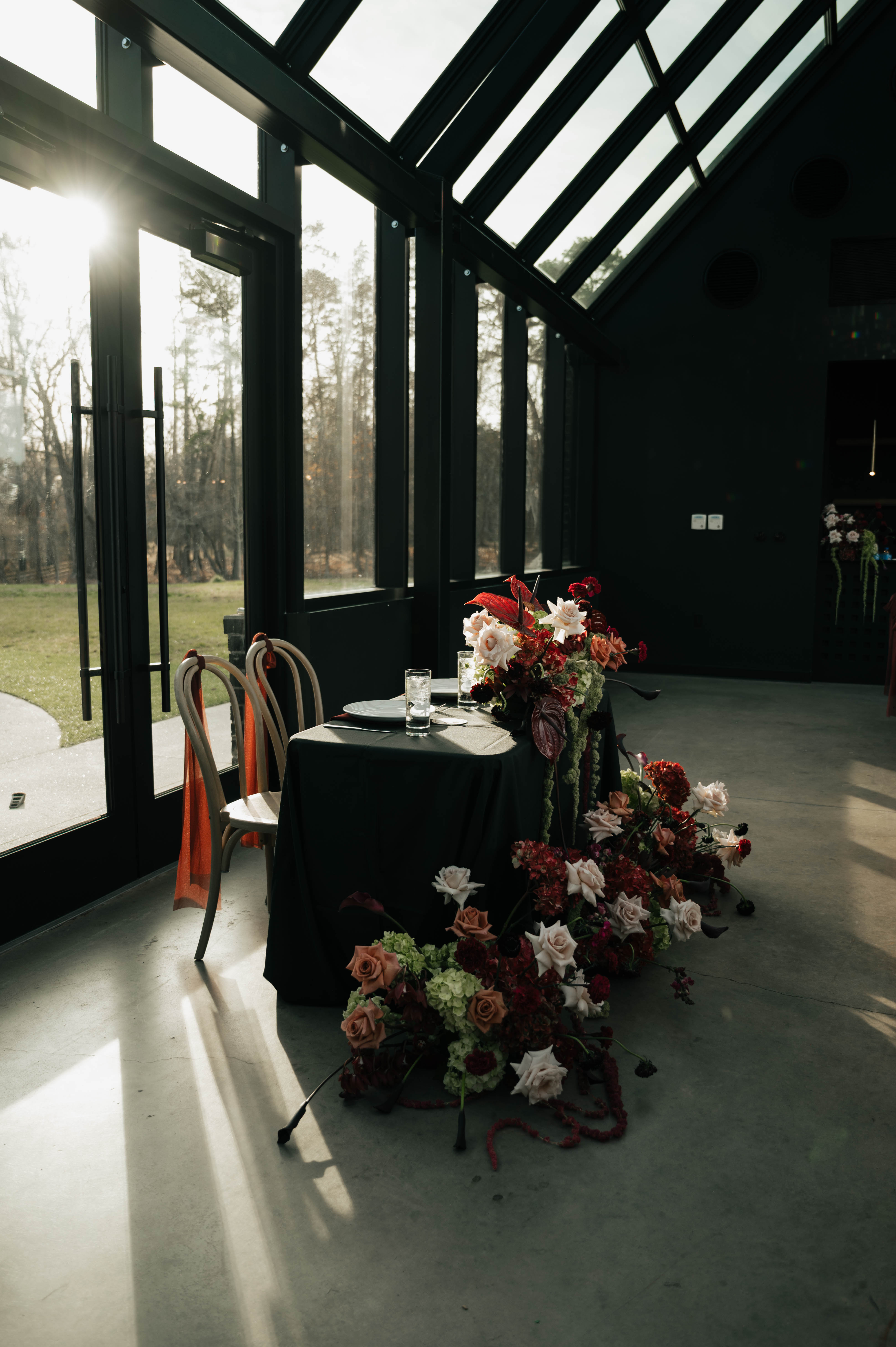 Dark greenhouse-style wedding venue with floor-to-ceiling windows and an oxblood floral arrangement on a pedestal, backlit by natural light