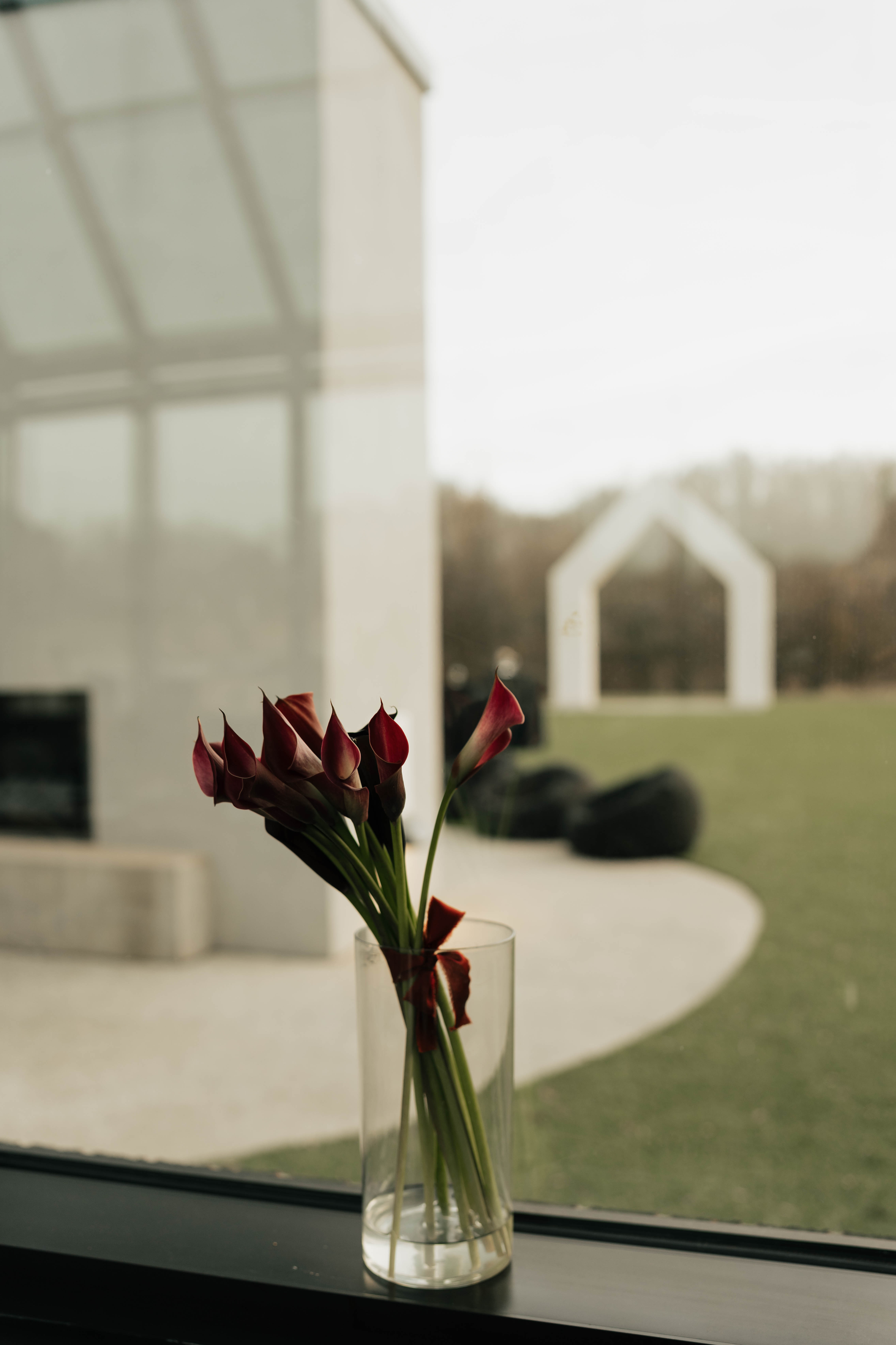 Small vase of deep red oxblood flowers on a windowsill in a modern glass wedding venue overlooking a lawn
