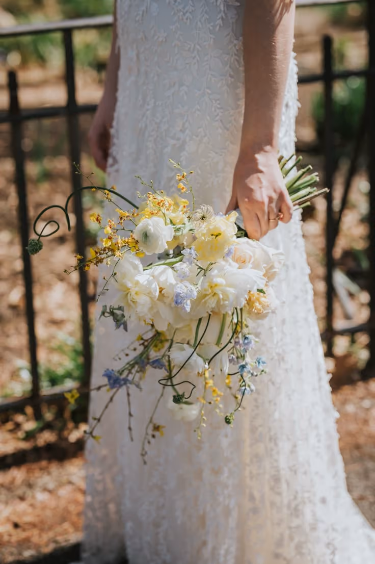 This may contain: a bride holding a bouquet of white and yellow flowers on her wedding day in front of a wrought iron fence