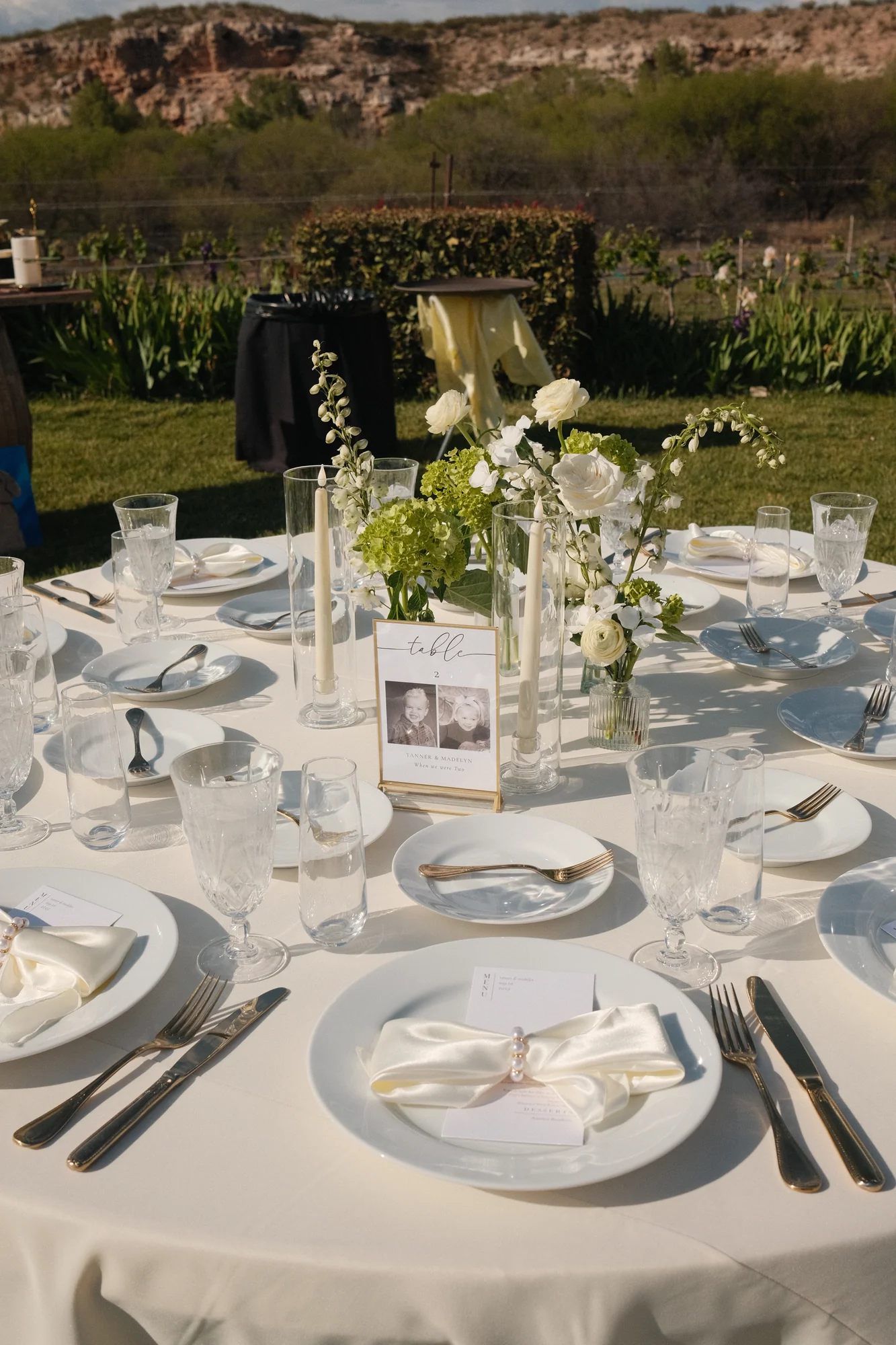 Close-up of a round outdoor reception table with white china, gold flatware, ivory ribbon-tied napkins, and a table number card with a vintage family photo, set against a rustic hillside backdrop