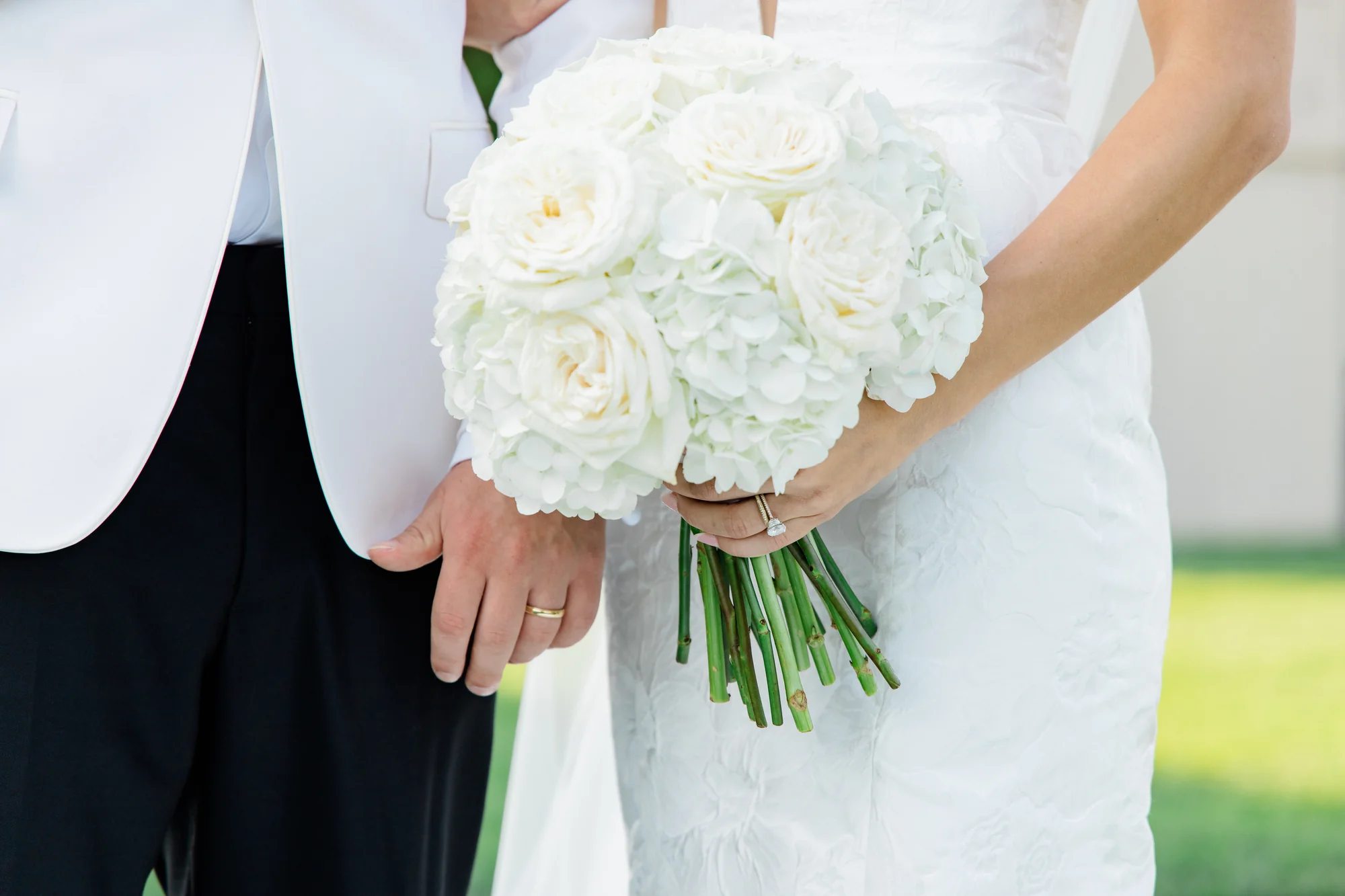 Bride holding a white bouquet of garden roses and hydrangeas next to groom in a white tuxedo jacket