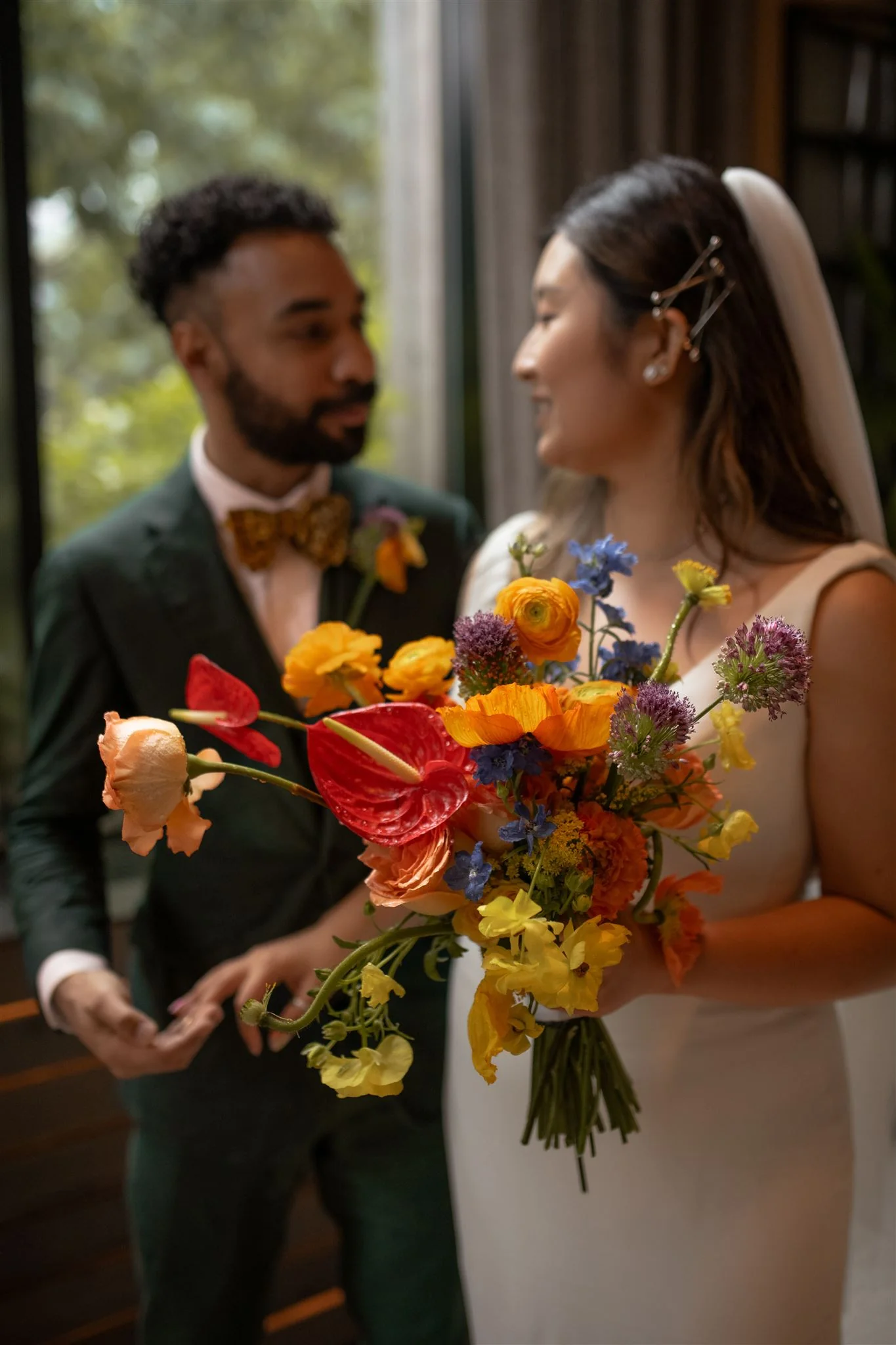 Bride and groom smiling at each other, bride holding a vibrant bouquet with red anthurium, orange poppies, yellow ranunculus, blue delphinium, and purple allium