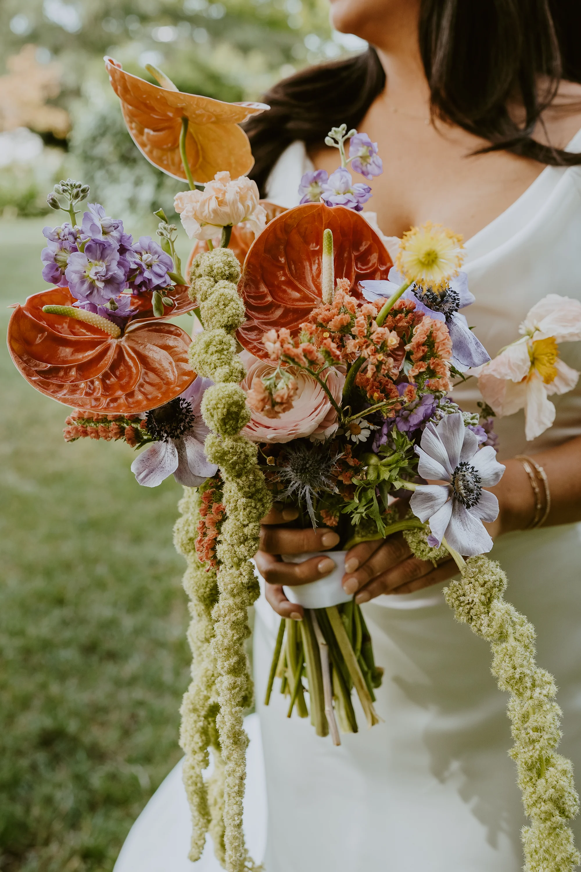 Close-up of a bouquet with orange anthurium, peach ranunculus, coral celosia, and purple anemones held by the bride