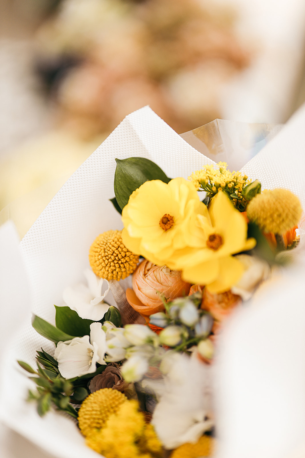 Close-up of a paper-wrapped bouquet with yellow ranunculus, yellow craspedia, peach roses, and white flowers