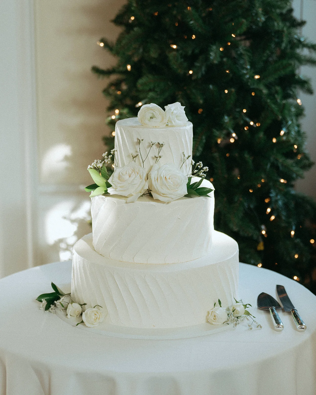 Close-up of a white tiered wedding cake decorated with greenery, near a lit Christmas tree