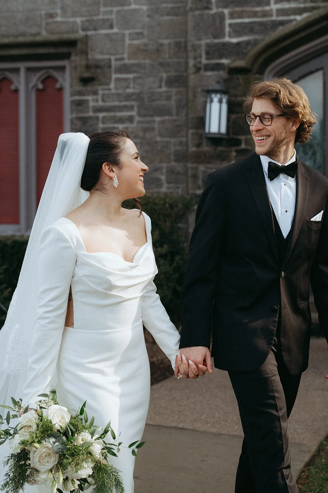 Bride and groom walking hand in hand in front of a stone building, the bride in a fitted long-sleeve white gown and the groom in a black tuxedo