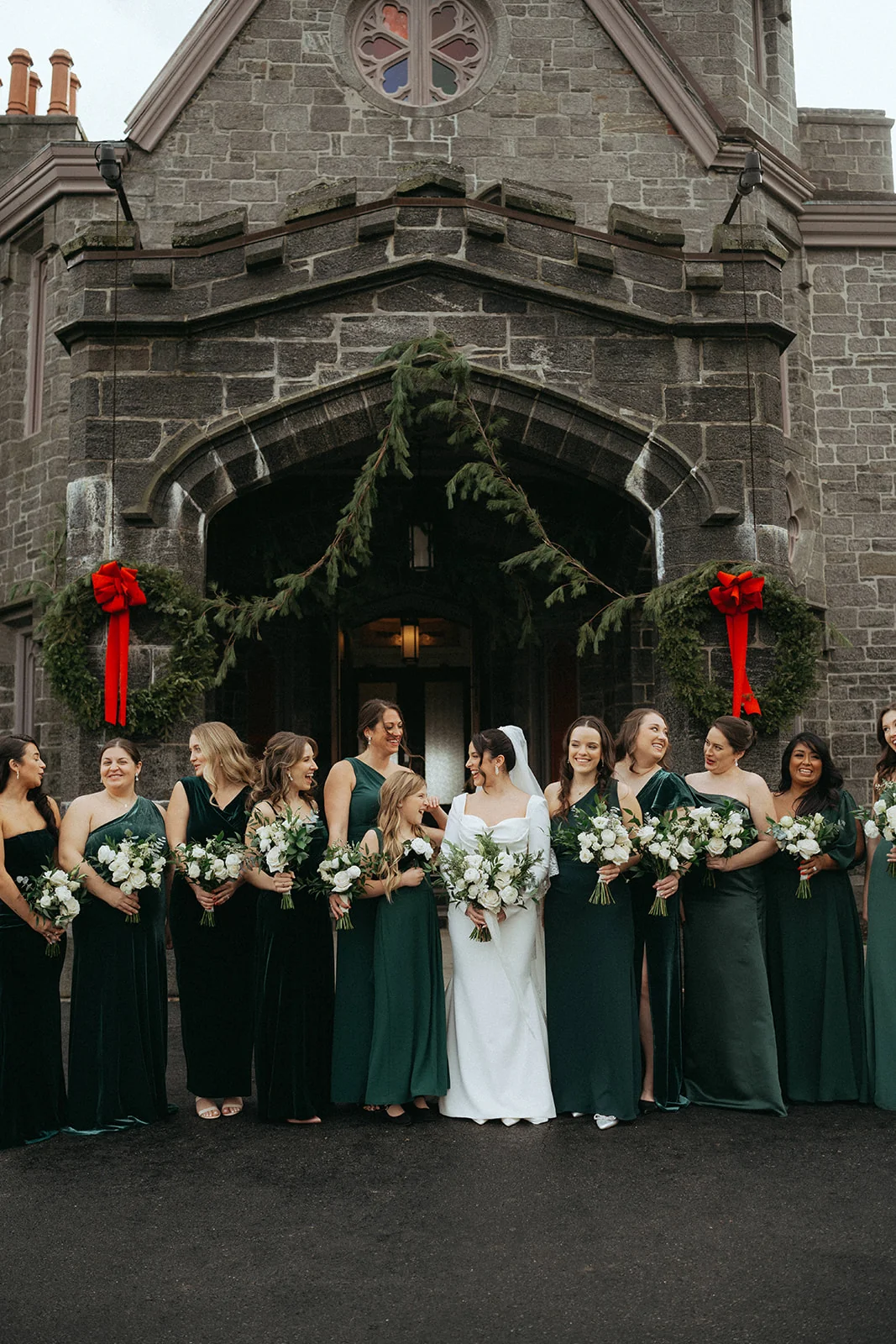 Wedding party in front of a stone church entrance decorated with red holiday bows, bridesmaids in dark green dresses and the bride in a white gown holding a white bouquet