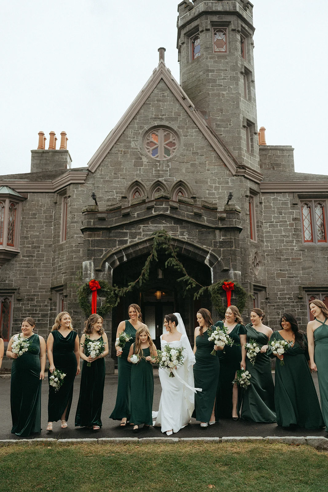 Wedding party with bridesmaids in dark green dresses and groomsmen in front of a grand stone building entrance decorated with red ribbon