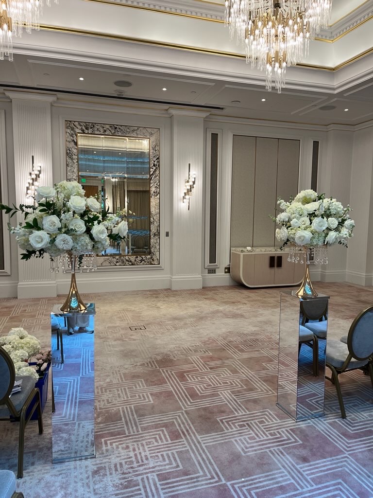 Close-up of white rose and greenery arrangements on mirrored pedestals at a wedding ceremony altar, with a large decorative mirror and chandelier in the background