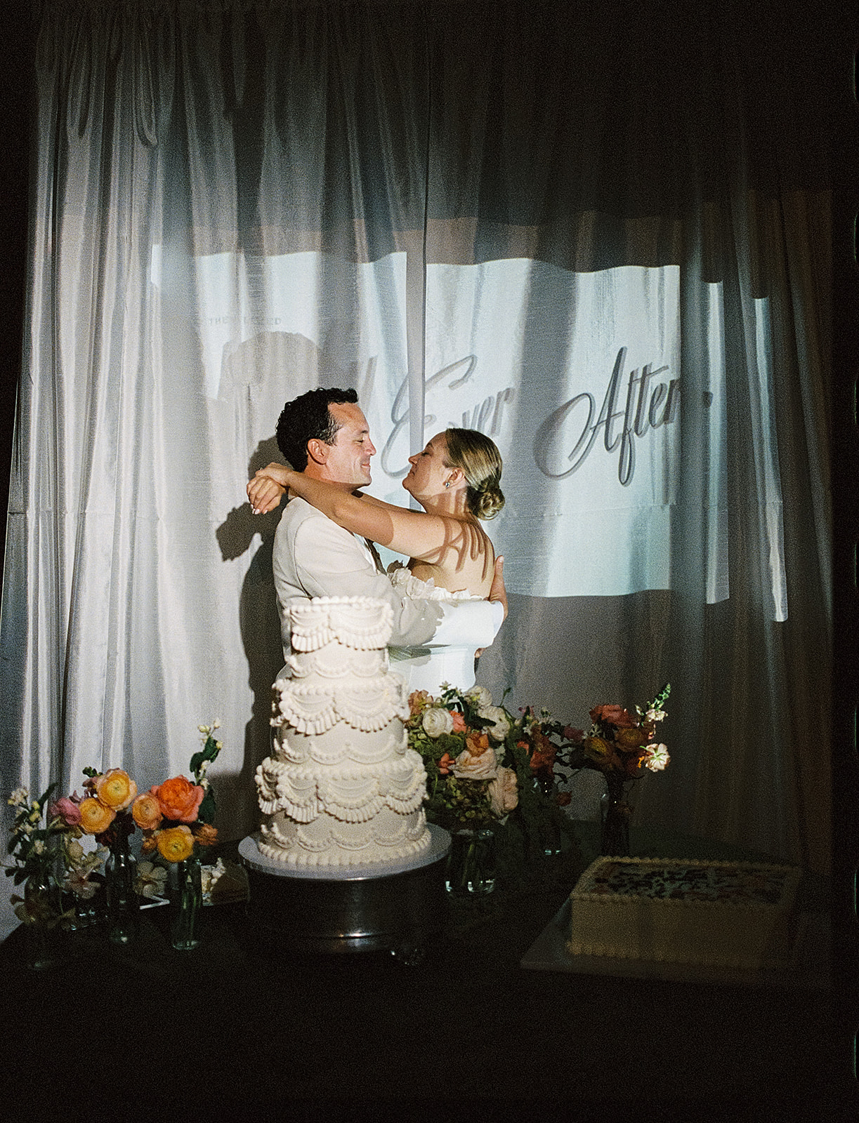 Vintage-style photo of a bride and groom embracing in front of a tiered wedding cake and "Ever After" backdrop, evoking an old-world, retro wedding portrait.