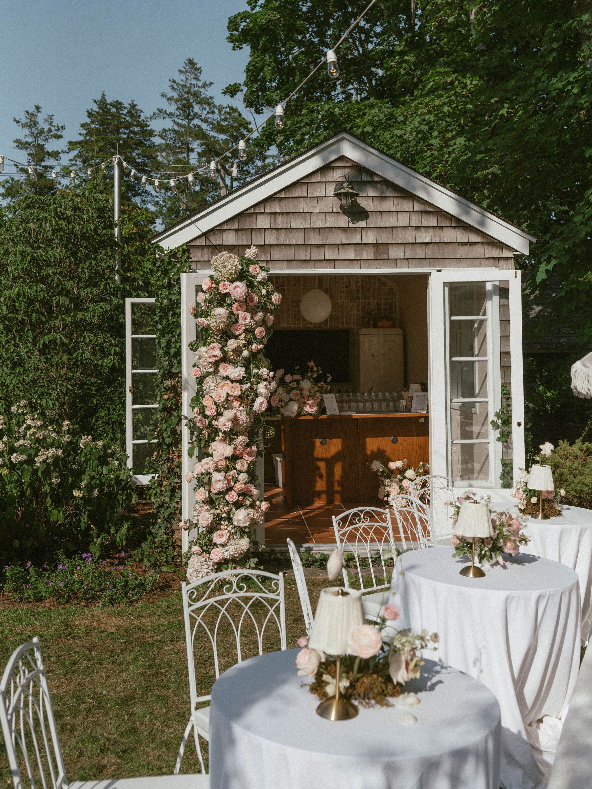 Small courthouse-style cottage entrance framed by a climbing pink rose archway, with intimate outdoor seating at round tables dressed in white linens and candlelight.