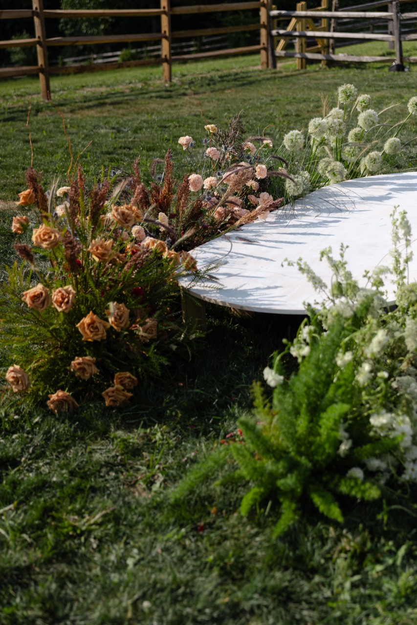 Loose, earthy wildflower arrangement of garden roses and textural grasses laid along a wooden fence line in an outdoor field setting.