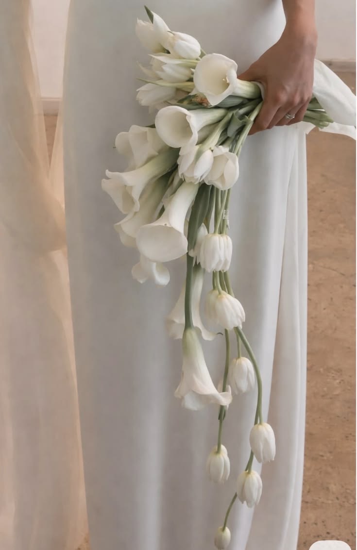 Bride holding a cascading bouquet of white calla lilies with long, exposed natural stems, set against a white wedding gown.