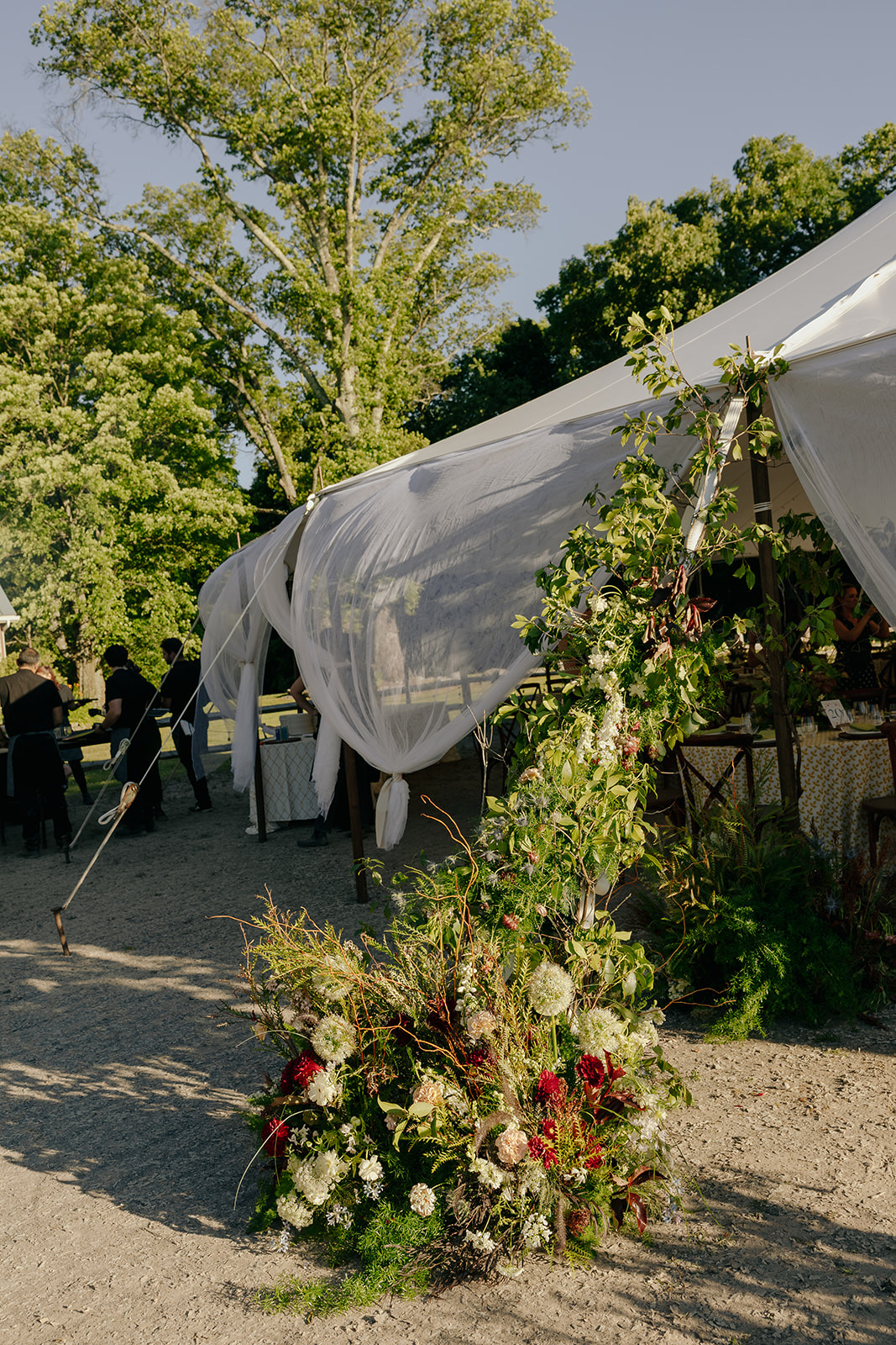 Tented outdoor wedding with a tall wild floral installation of red blooms, greenery and grasses climbing the tent entrance