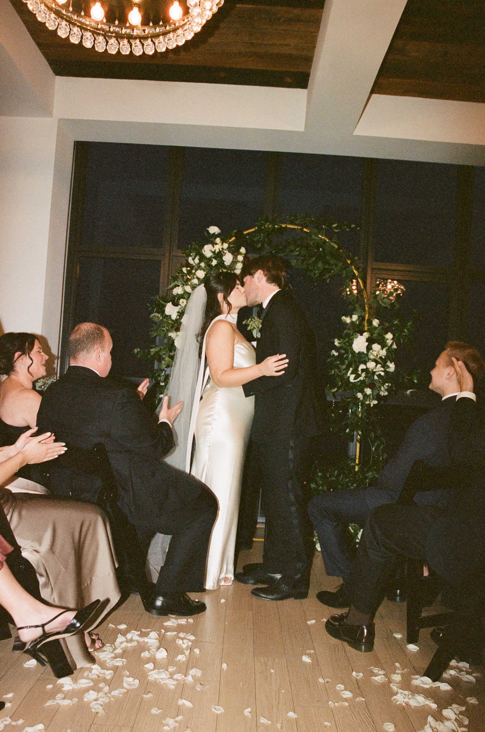 Couple kissing at an intimate indoor wedding ceremony in front of a floral arch, with flower petals scattered on the floor