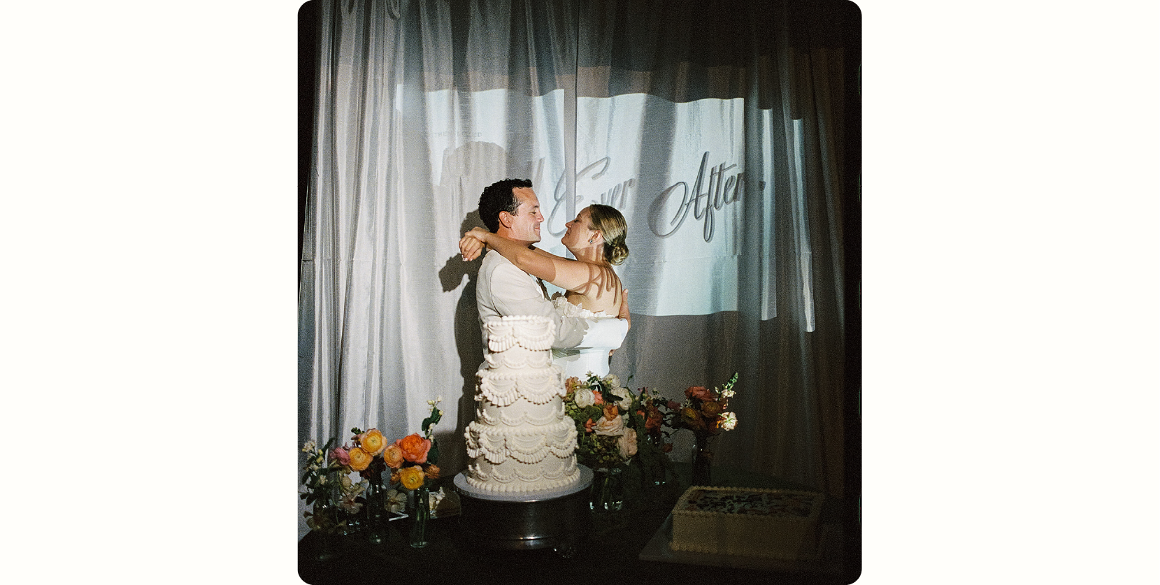 Nostalgic wedding trend for 2027: a couple embracing beside a tiered white lace wedding cake with warm orange and peach florals, shot on film in front of sheer draped curtains