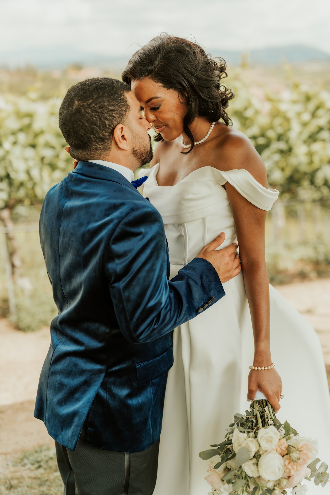 Groom in a navy velvet tuxedo kissing the bride, who holds a blush and white rose bouquet, at Callaway Vineyard & Winery