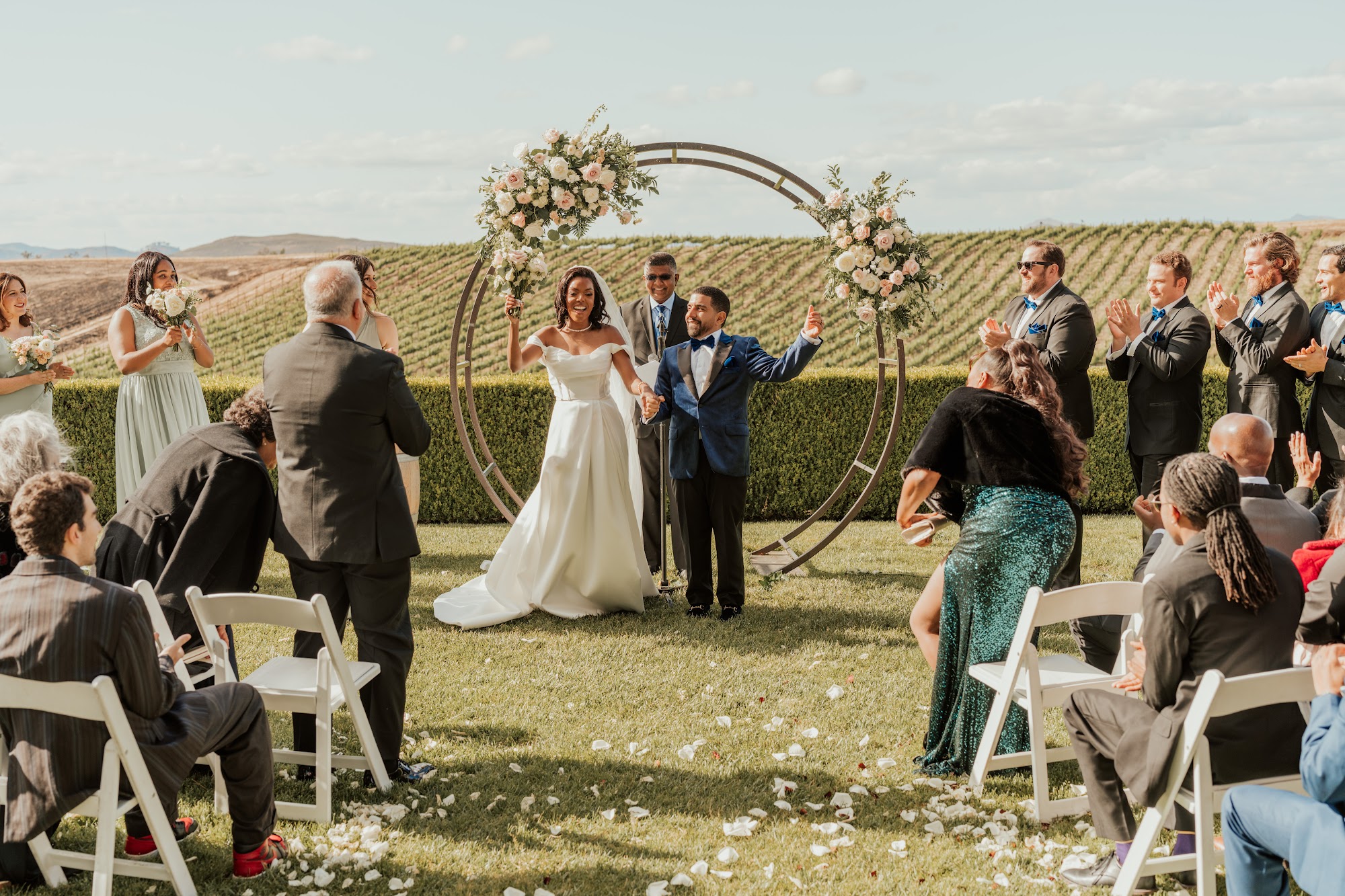 Guests cheering as the couple celebrates under a blush and white floral circular arch at an outdoor Temecula vineyard ceremony