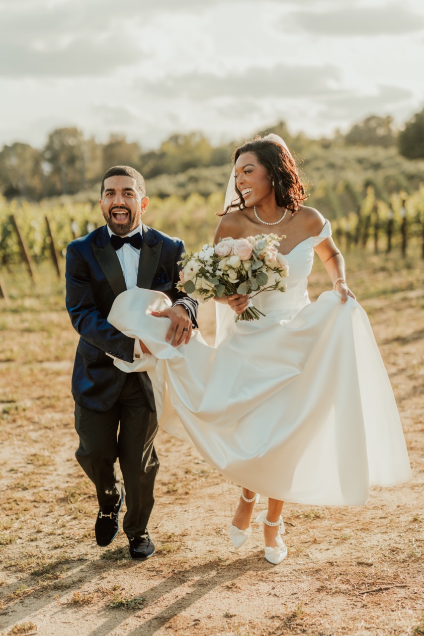 Bride with a blush and white rose bouquet and groom in a navy tuxedo laughing as they walk through the vineyard at Callaway Vineyard & Winery