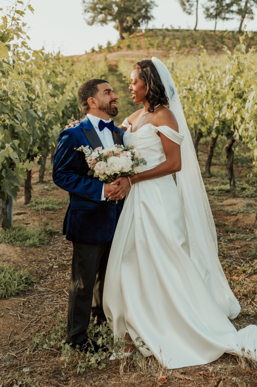 Bride and groom embracing among the grapevines at a Temecula vineyard wedding, bride holding a blush and white rose bouquet
