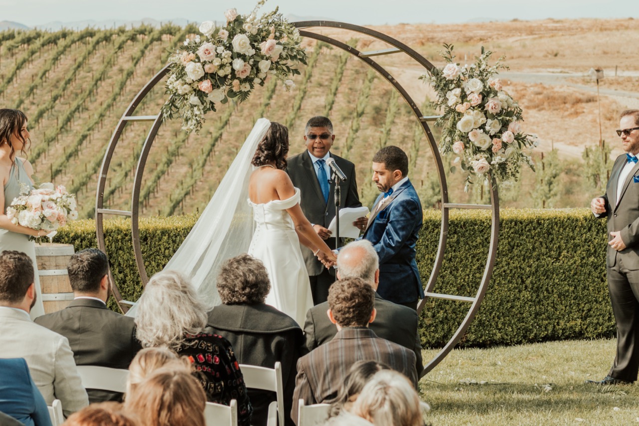 Bride and groom at the altar under a circular arch with blush and white flower clusters overlooking Temecula vineyard rows