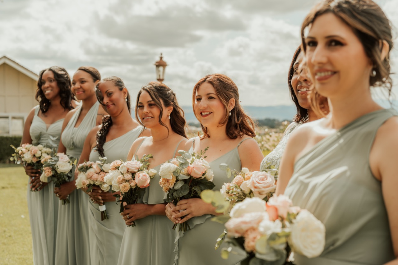 Bridesmaids in sage green dresses holding blush and white rose bouquets at a Temecula vineyard wedding ceremony