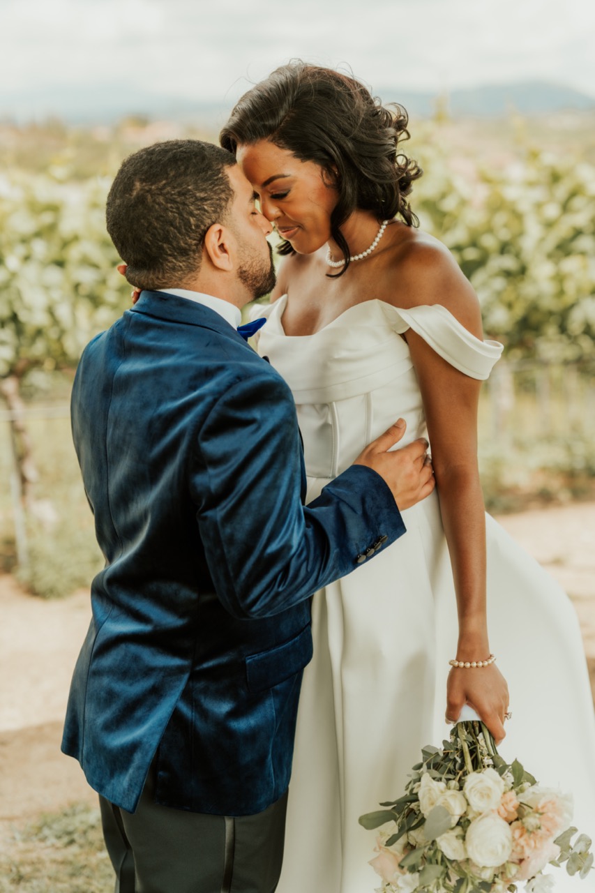 Groom in a navy velvet tuxedo kissing the bride, who holds a blush and white rose bouquet, at Callaway Vineyard & Winery