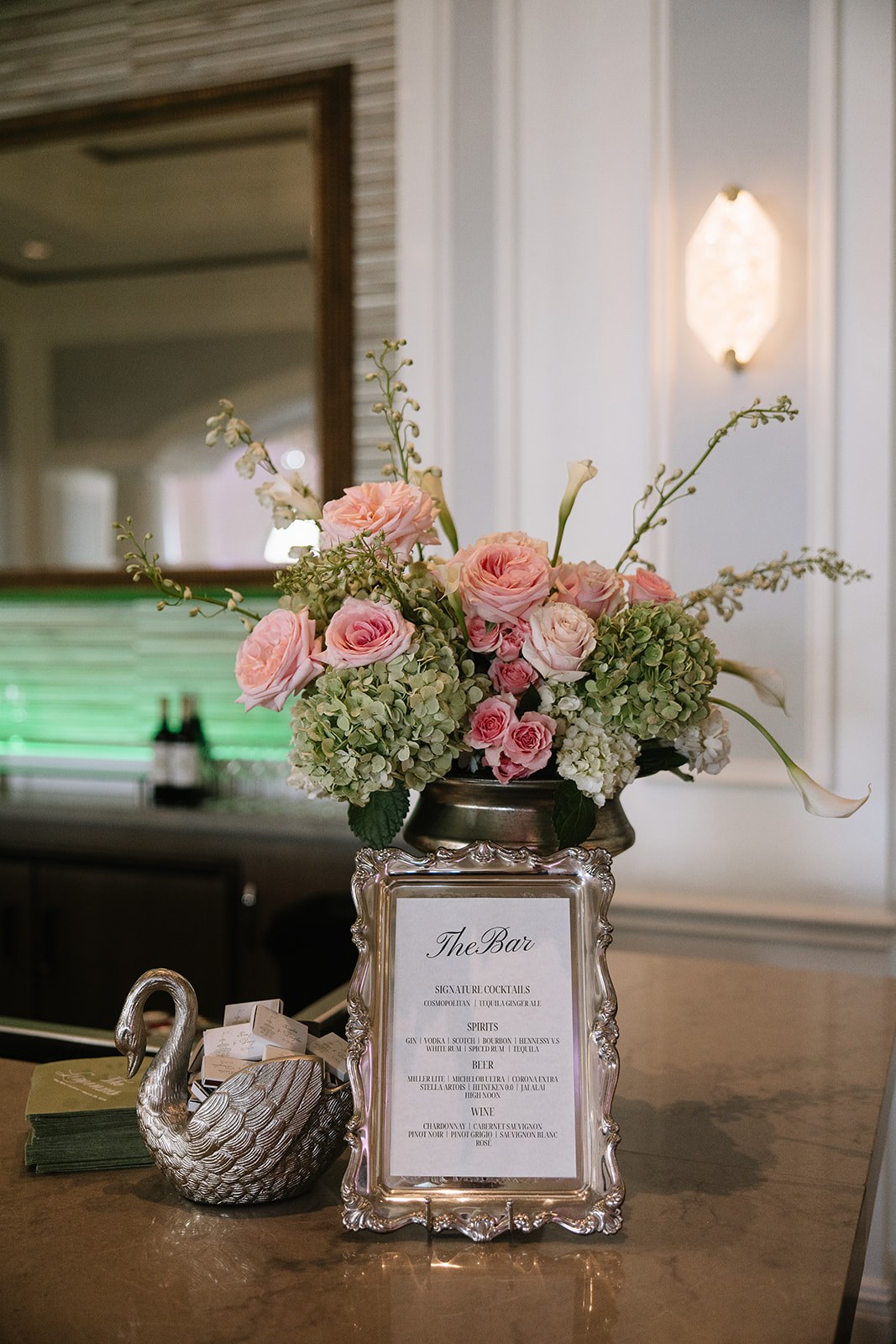 Bar arrangement of green hydrangea with pink garden roses, spray roses, curving white calla lilies and delphinium in a silver urn
