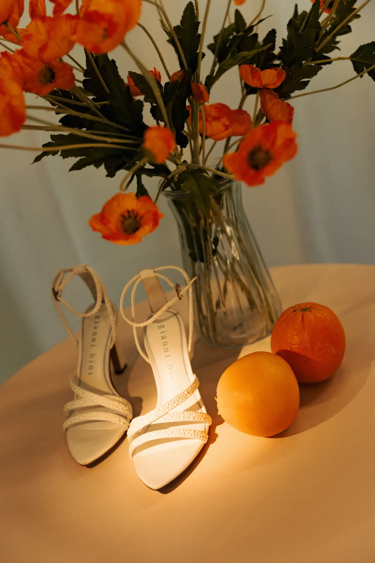 Orange flowers in a glass vase beside white wedding shoes and oranges