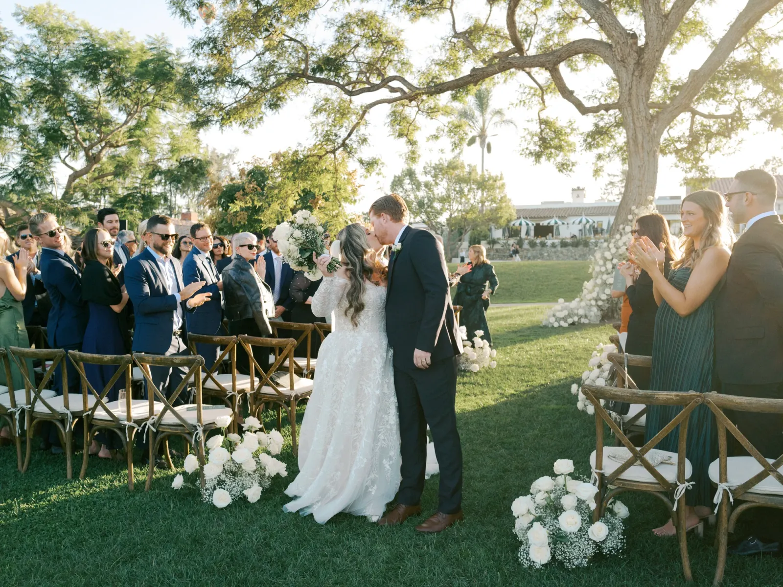 A couple at the end of a golden-hour ceremony aisle lined with white roses and baby's breath