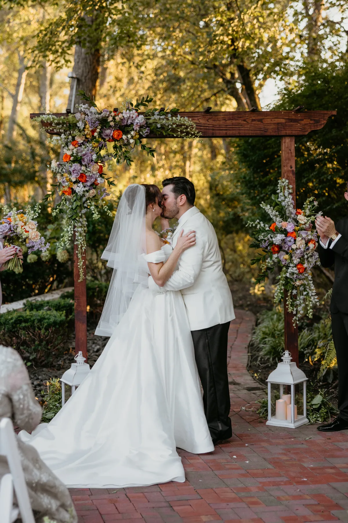 A couple kissing under a wooden ceremony arch dressed with orange ranunculus and dusty lavender roses