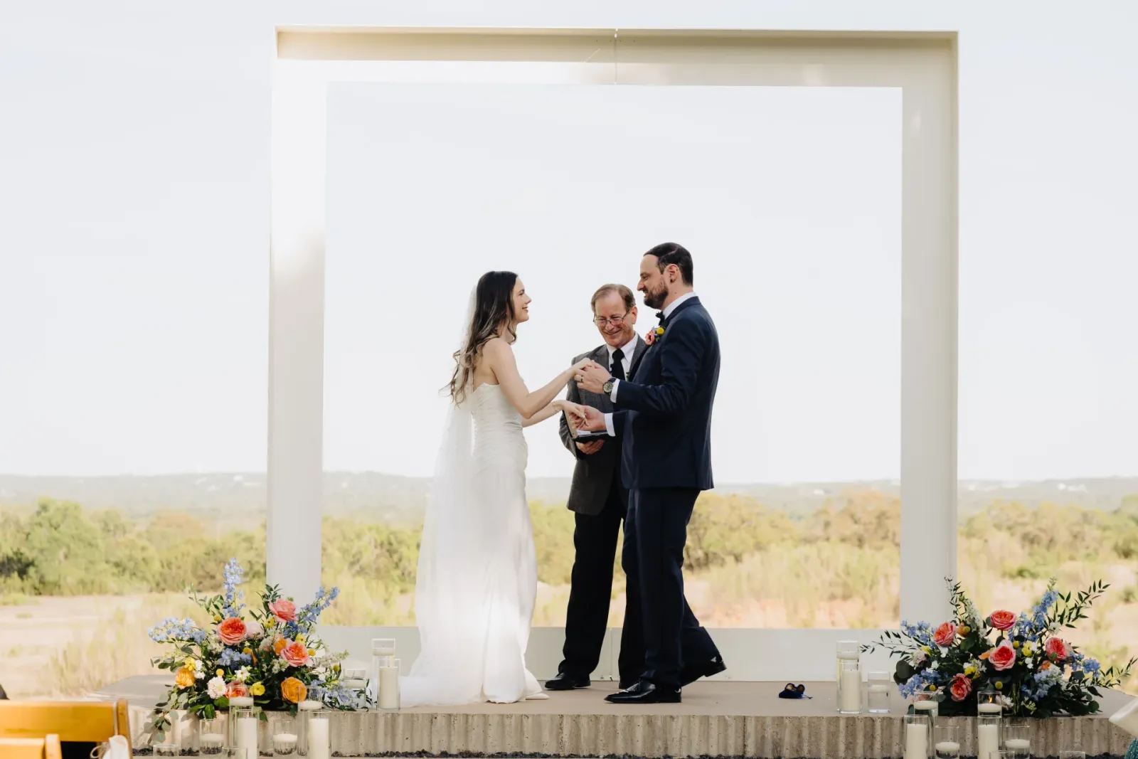 Ceremony flowers of light blue delphinium, coral roses and yellow craspedia framing a couple at the altar