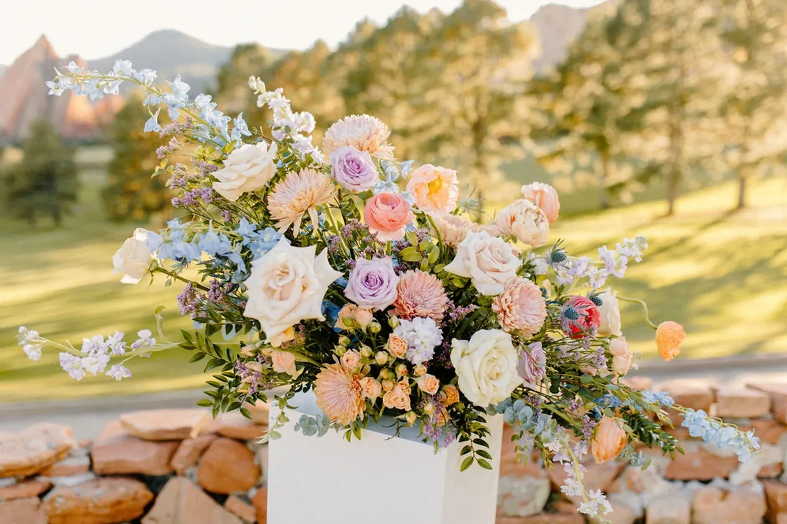 A Poppy arrangement of peach and lavender roses with blue delphinium on a stone wall at sunset