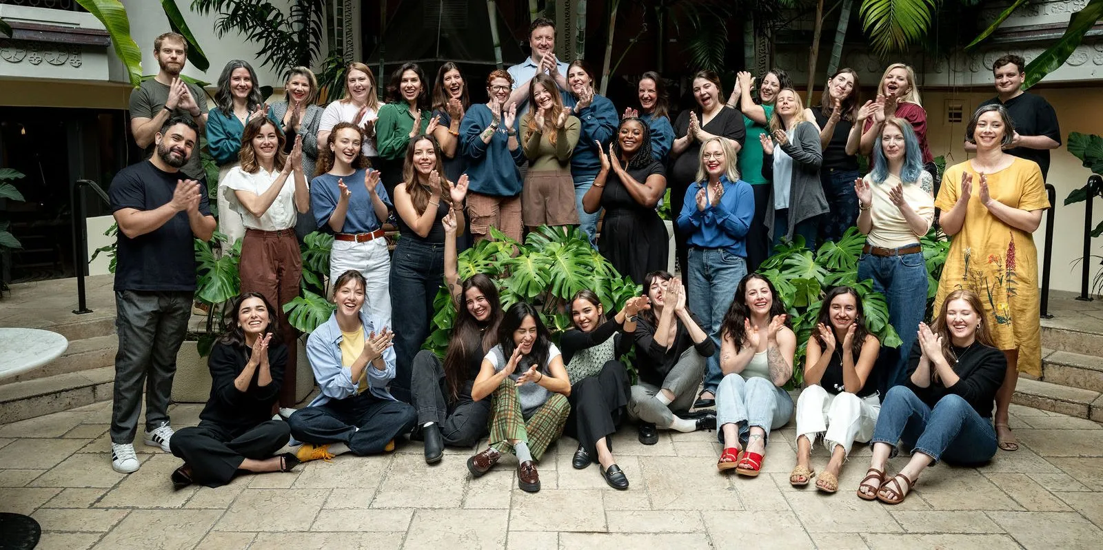 The Poppy team, about forty people, clapping and laughing together in a courtyard full of palms and monstera
