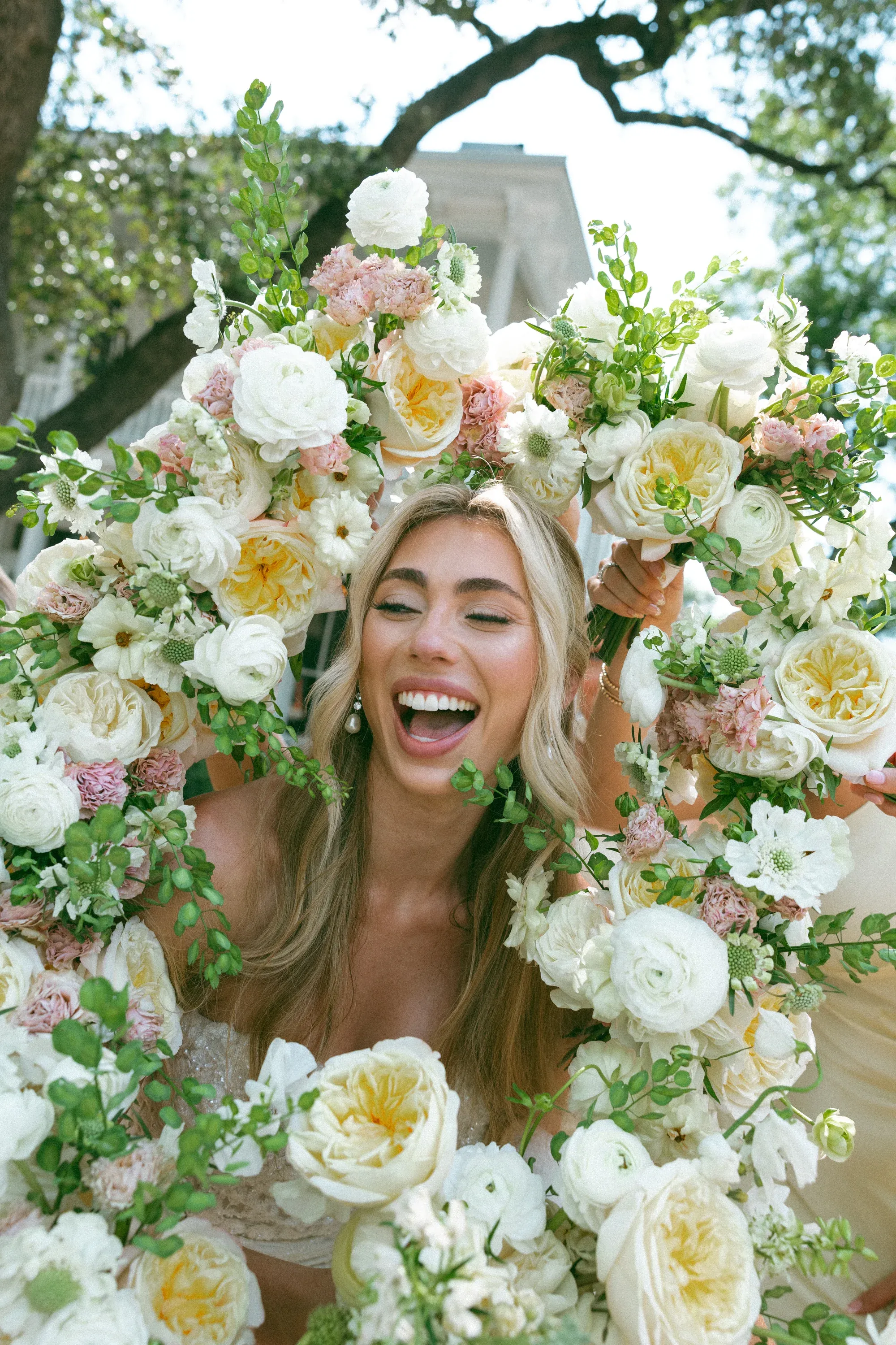 A laughing bride framed by blush and ivory garden-rose bouquets her bridesmaids hold up around her