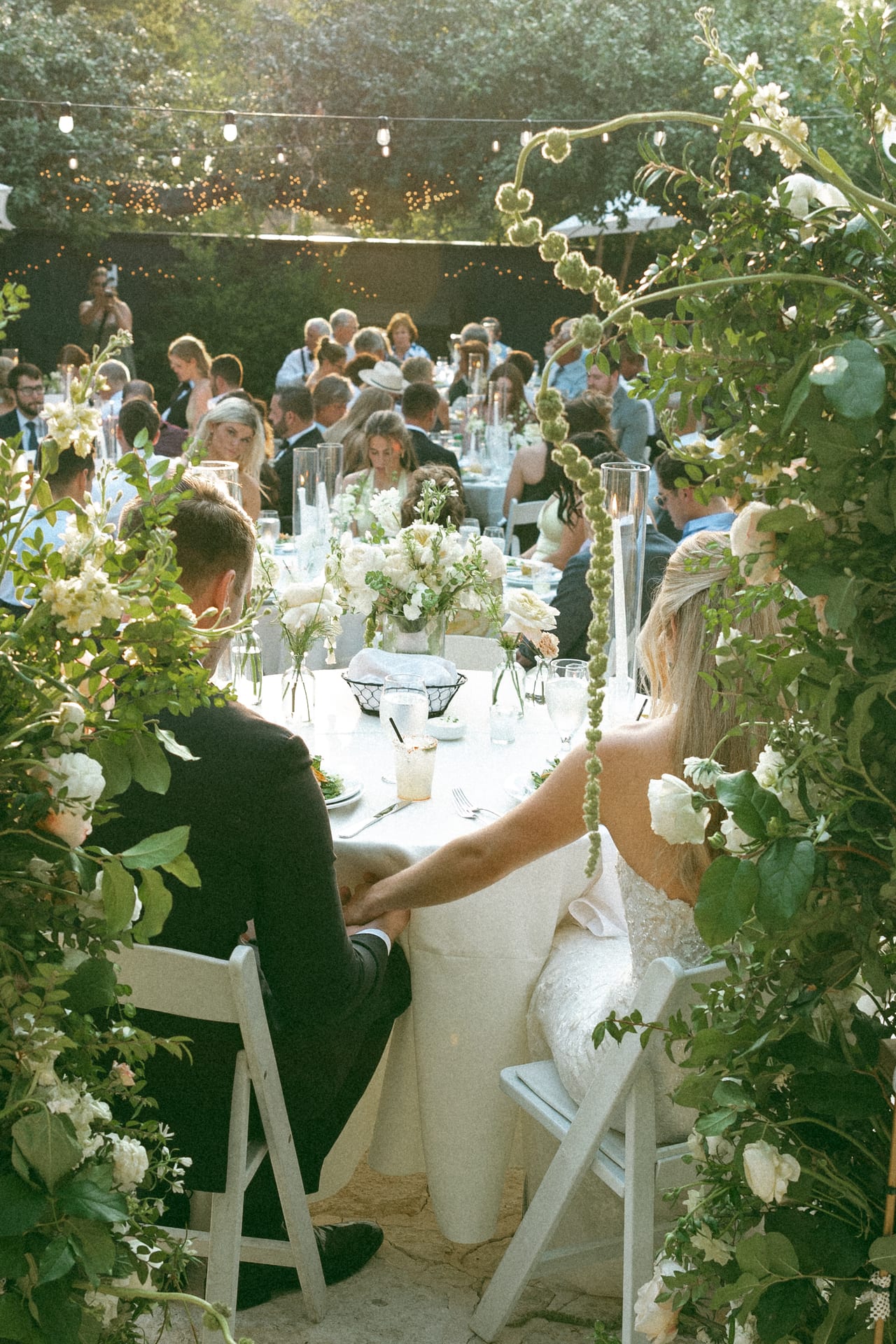 Bride and groom seated beneath abundant greenery and white floral installations at an outdoor wedding reception.