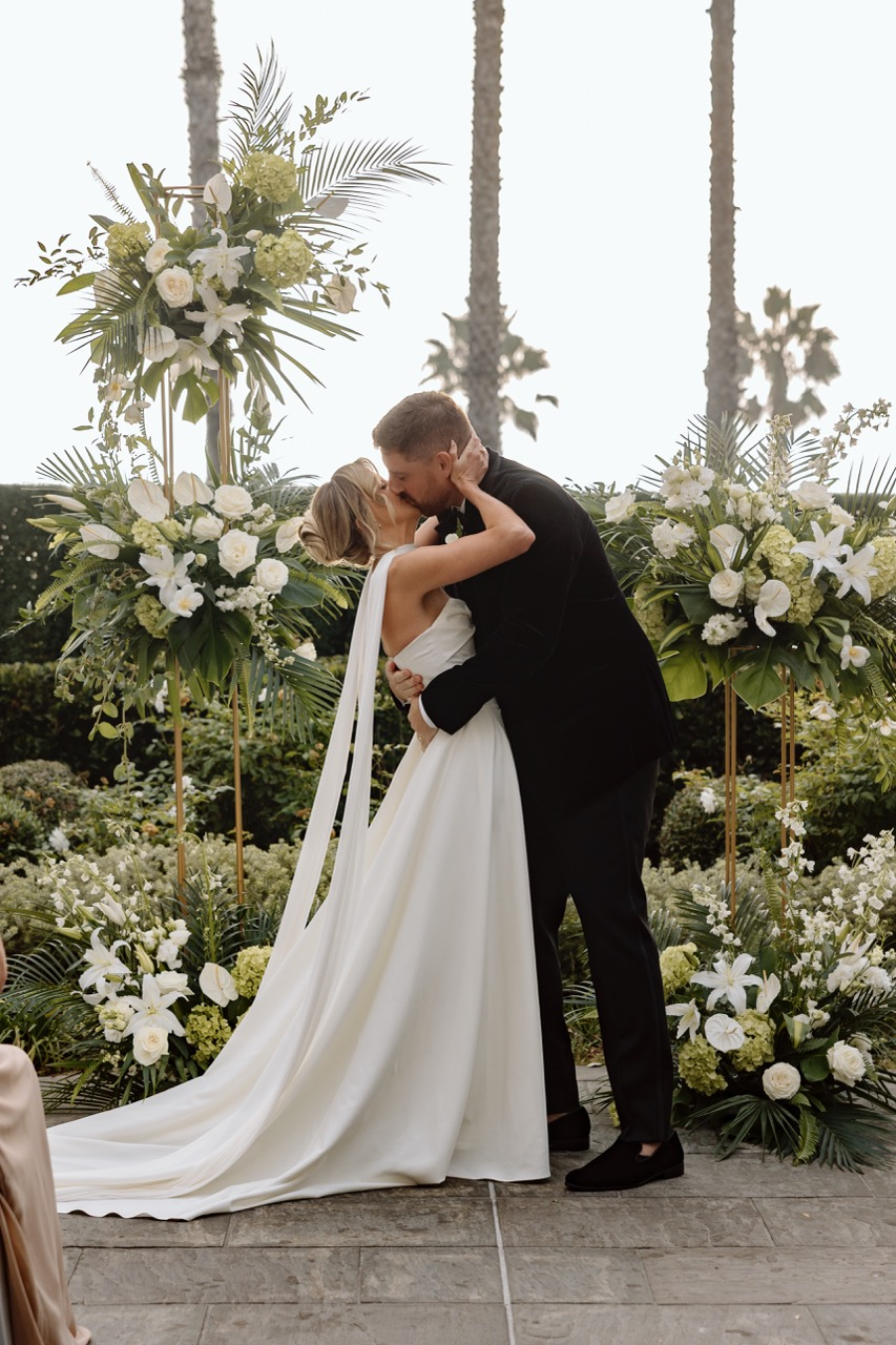 Newlywed couple kissing beneath white and green tropical ceremony florals at The Seabird Ocean Resort