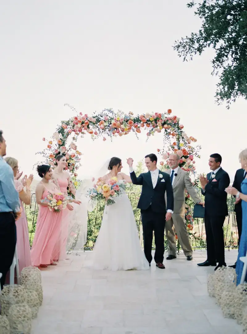 Wedding couple walking beneath a colorful floral arch at Villa Antonia
